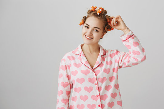 Indoor shot of positive cosy woman wearing curlers and lovely pyjamas with heart print, touching waverer and smiling at camera, standing over gray background. Girl wants to have great haircut tomorrow