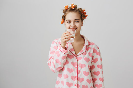 Water makes you beautiful. Indoor shot of attractive caucasian girl in hair curlers and nightwear with heart template drinking glass of water and smiling broadly, standing over gray background.