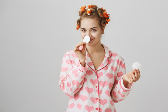 Shall we do some beauty today. Studio shot of interesting cute girl in hair curlers and nightwear, holding cotton pads in hands and covering nose, smiling intriguing over gray background