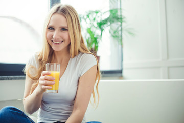 Smiling beautiful young blond woman sitting at home on the couch with a glass of orange fresh.