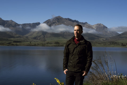 Retrato de hombre joven frente a paisaje de montañas con picos nevados y nubes. Las montañas se reflejan en un lago. Escena diurna, cielo azul y despejado. Nueva Zelanda.