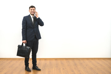 The happy businessman with a briefcase phones on the white wall background