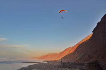 The paraglider flies over the coast at sunset. Red rocky coast of Legzira beach, Morocco.