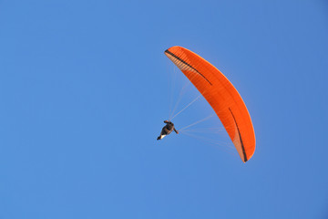 One person safely flies on a paraglider with an orange wing in the evening against a blue sky.