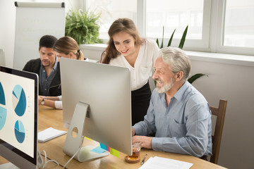 Smiling young manager helping senior worker with funny computer work in office, mentor teacher...