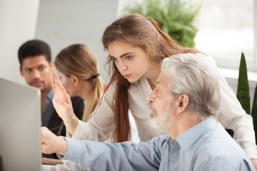 Older employee asking young female manager question about online task, corporate teacher helps aged office worker with computer work explaining senior businessman how to use new corporate application