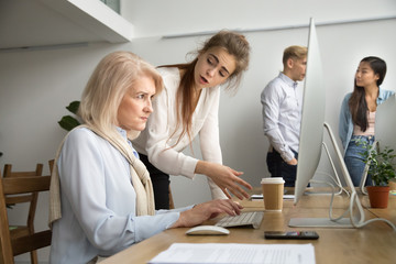 Young team leader correcting offended senior employee working on computer in office, female manager...