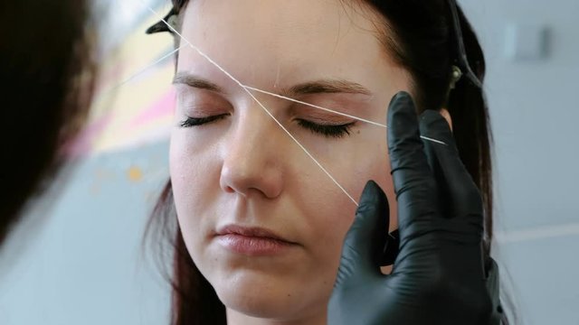 Cosmetologist performs the procedure of correction eyebrow threading. Front view.