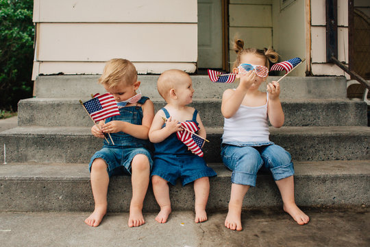 Blonde toddler siblings wearing denim waving American flags outside in the summer.