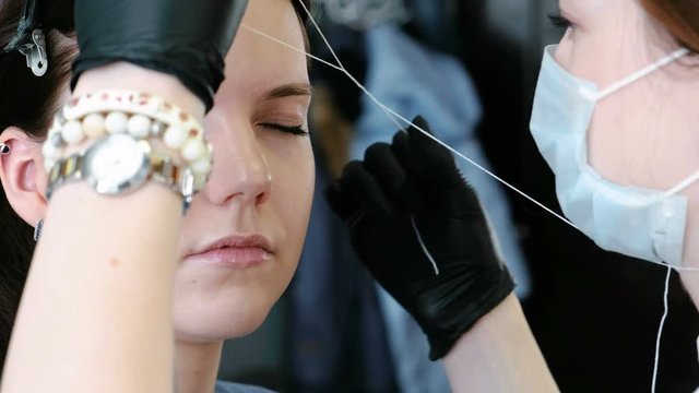 Cosmetologist performs the procedure of correction eyebrow threading. Side view.