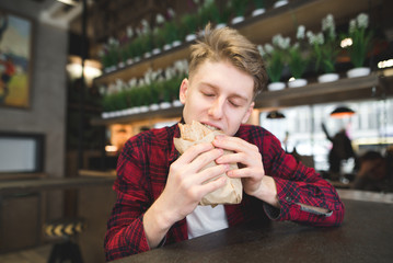 A handsome young man with pleasure bites an appetizing sandwich in a cafe. A student dishes a sandwich in a cafe