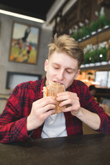 A young man bites a panini sandwich in a cafe. The student is sitting in a cozy cafe and dining with a sandwich