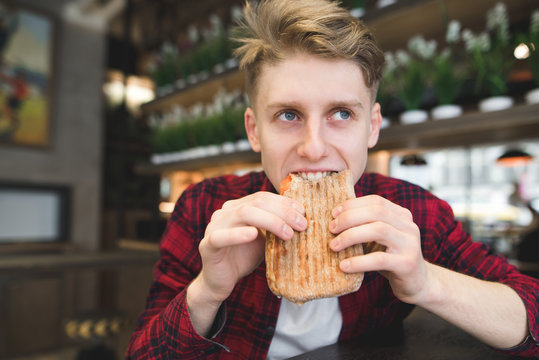 A beautiful young man eats a panini sandwich in a cozy cafe. Funny student bites sandwich. Lunch at the cafe.