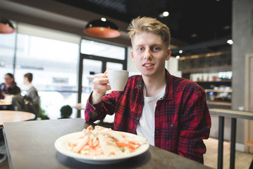Happy young man sits in a restaurant with a plate in a plate and a cup of coffee in his hands. The student dishes in the cafe. A look at the camera