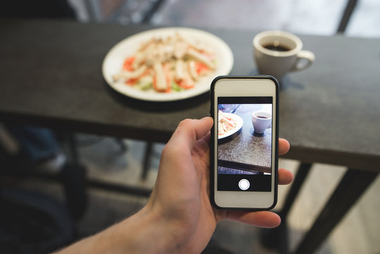 The hand with the phone makes a photo of the food in the restaurant. A salad and coffee photo on a smartphone