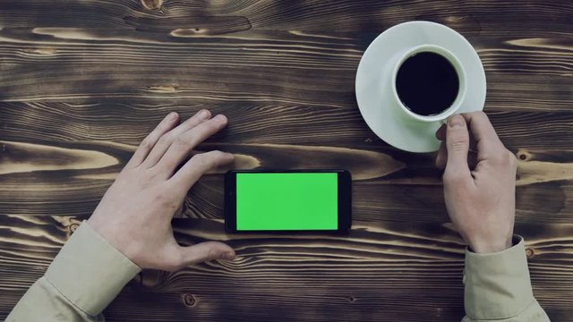 White cup coffee and smartphone with green screen in horizontal position on wooden table. Hands of young man in shirt. He drinks coffee, looks and scrolls finger to right on left of menu mobile app.