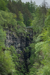 Fototapeta premium CBraemore, Scotland - June 8, 2012: Corrieshalloch Gorge, a deep cut in landscape with forested vertical slopes. Suspension bridge over chasm. Focus on black rocks and abundance of green trees.