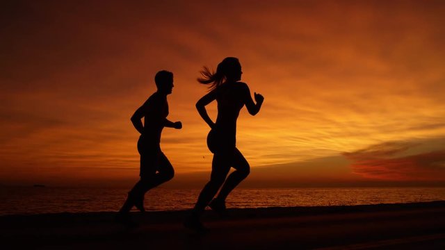 SILHOUETTE: Young couple having fun on tranquil run at beautiful orange sunset. Unrecognizable active woman and man bond through jogging down ocean road on a breathtaking summer evening at seaside.