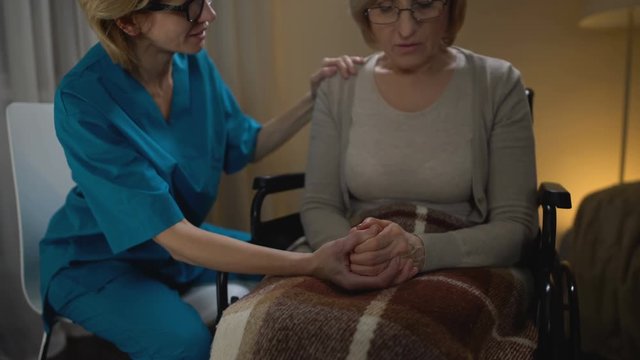 Nurse talking heartily to old woman in wheelchair supporting her, elderly home