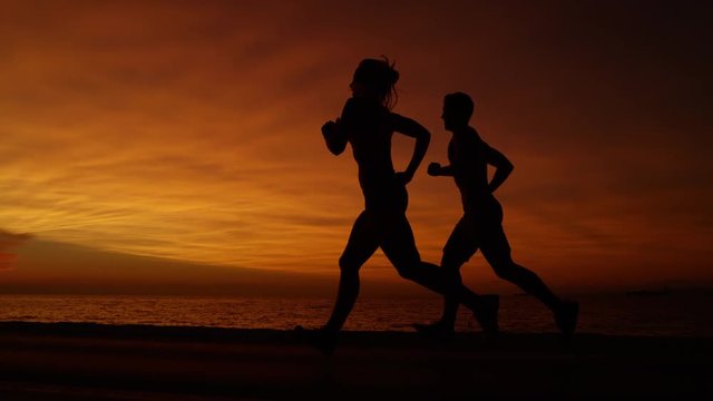 SLOW MOTION, SILHOUETTE: Young woman and man go for an evening jog on a breathtaking evening at the seaside. Awesome slow motion shot of couple jogging along the sea in front of stunning orange sunset