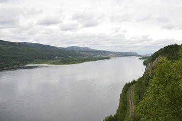 Fototapeta premium The river flows between hills and forests, alongside a highway and a railway line. Gloomy, overcast sky with clouds.