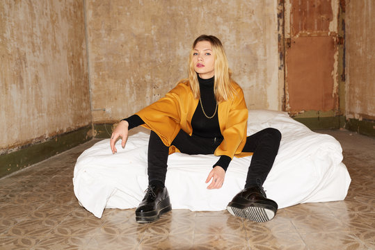 Pretty blonde girl in yellow coat sitting on mattress in shabby