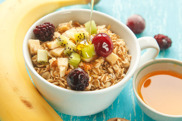 Cup of oat flakes with cherry, kiwi, blackberry, egg, walnut and honey in small cup on a turquoise wooden table.
