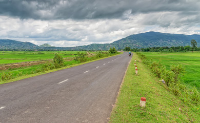 Fototapeta premium asphalt road, among the fields, with people riding a moped towards the mountains, against the background of clouds, sky and green vegetation
