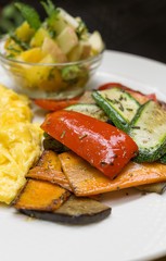 Baked vegetables on white plate: sliced carrots, bell pepper, zucchini with scrambled eggs and blurred mango apple salad in background. Vegetarian food concept. Detox