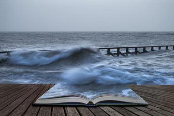 Beautiful dramatic stormy landscape image of waves crashing onto beach at sunrise in pages of book