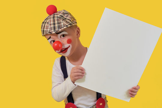 Funny child clown peeks out blank white paper. little boy clown in makeup,red nose. Little cheerful boy jester keep sheet with copy space for your text. Concept birthday, holiday,celebration, party.
