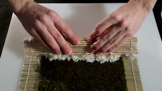 A man with his bare hands makes a roll with a shrimp. On the table is a sheet of nori, a bamboo mat, rice and shrimp. Close-up