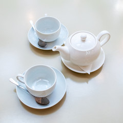 Two white ceramic tea mugs with shiny spoons and saucers and a teapot stand on the table in a cafe