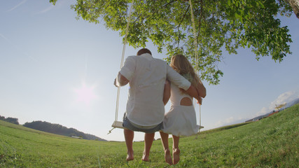 CLOSE UP: Carefree young couple swaying on wooden rope swing on sunny summer day