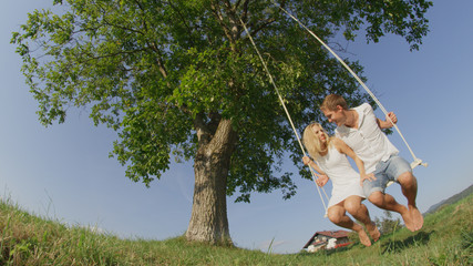 CLOSE UP LOW ANGLE: Lovely infatuated couple swaying on a swing in green meadow.