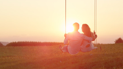 LENS FLARE: Happy couple swaying on a swing embraced on sunlit summer evening.