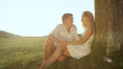 Fototapeta na wymiar CLOSE UP: Embraced young couple in love sitting under a beautiful tree in meadow