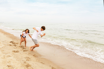 Two teenagers: a girl and a boy with blond hair, dressed in white T-shirts they run, laugh and play in the sea beach.