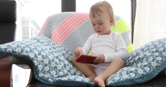 Adorable toddler boy sitting on a chair and playing with smartphone. Child learning how to use smartphone. Technology and lifestyle concept