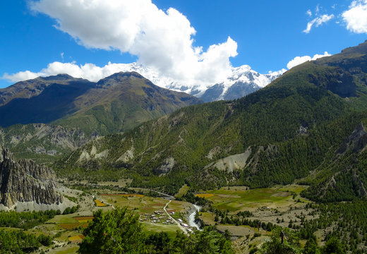 A village beside a river below green sided mountains and snow covered mountain tops