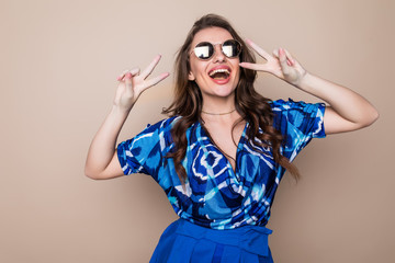 Portrait of a smiling happy playfull woman in sunglasses showing victory sign and looking at camera isolated on the color background