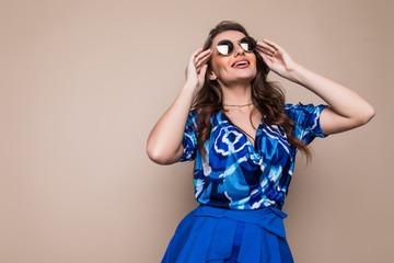 Portrait of a joyful young woman dressed in blue dress and sunglasses looking at camera isolated over brown background
