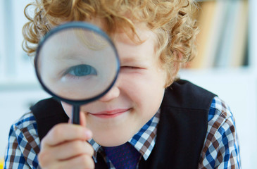 Little Caucasian boy looking at camera through magnifying glass in classroom. Studying the microworld with tools.