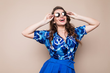 Portrait of a joyful young woman dressed in blue dress and sunglasses looking at camera isolated over brown background