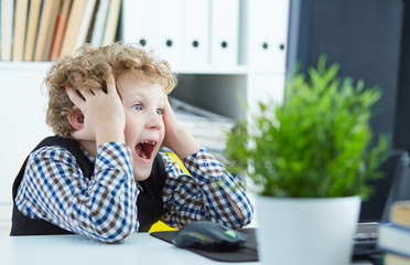 Little Caucasian curly boy looks at the computer screen and screams, clutching at the head