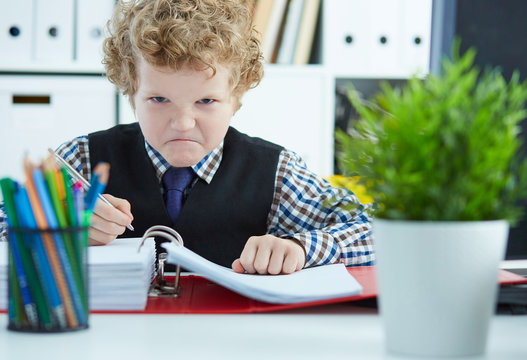 Little angry curly Caucasian boy like businessman doing paperwork in office.