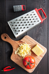 Grater, tomato and cutting board with grated cheese on wooden background