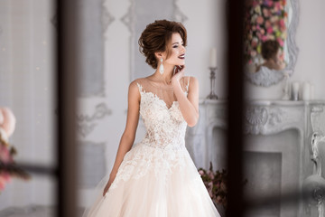 Morning of the bride. A beautiful bride is standing in a lace wedding dress. Wedding hairstyle and makeup. Hand at the face. Photo through the window.