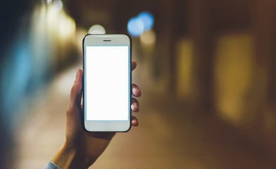 hipster using in hands clean gadget mobile phone closeup, blogger pointing finger on blank screen smartphone on background bokeh light in night atmospheric city, mockup online wifi internet concept