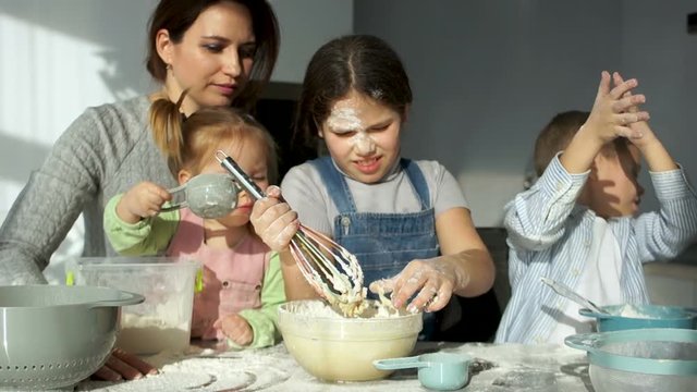 A beautiful mother with three cheerful children of different ages kneads the dough. The elder daughter removes the dough from the corolla, the younger sister helps knead the dough